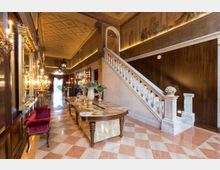 Interno di un palazzo storico con ampio atrio illuminato da lampadari, pavimento in marmo a scacchiera e soffitto decorato. Una scala in pietra con balaustra bianca sale lungo una parete in legno, accanto a un lungo tavolo con vasi e specchi dorati.