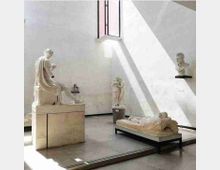Sala museale luminosa con pareti bianche e grande finestra alta che proietta fasci di luce diagonali sul pavimento. Sono esposte sculture in marmo su piedistalli e una figura distesa su una piattaforma, con un busto fissato alla parete.