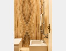 Bagno con parete rivestita in lastre di pietra venata a libro in tonalità beige e marrone, che crea un motivo simmetrico centrale. In primo piano si vedono una vasca bianca, una mensola/struttura bianca e un portasciugamani in metallo cromato con rubinetteria a vista.