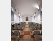 Interno di una ex chiesa adibita a sala conferenze, con file di sedie grigie disposte su due blocchi e corridoio centrale su pavimento a riquadri. Pareti bianche con soffitto a volte, finestre alte con tende e illuminazione a parete; in fondo un altare con nicchia e statua.