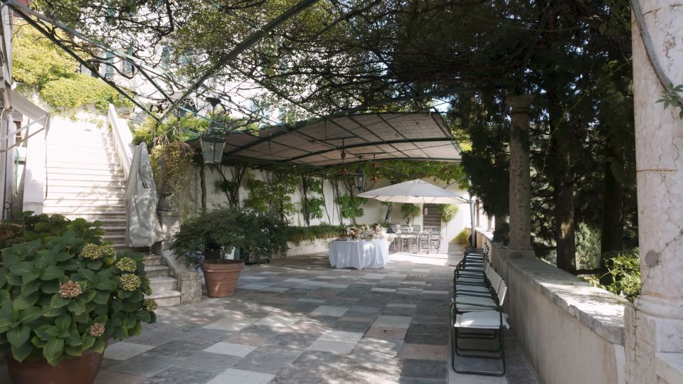 Terrazza in pietra con pavimentazione a lastre, coperta da una pergola con rampicanti e circondata da alberi. Sul fondo ci sono un tavolo apparecchiato sotto un grande ombrellone bianco e sedie allineate lungo un muretto; a sinistra una scalinata in muratura sale verso l’edificio.