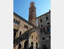 Facciata di un edificio storico in pietra a fasce bianche e rosse con arcate e finestre ad arco, affiancato da altri palazzi. Sullo sfondo svetta un alto campanile in mattoni con logge e coronamento superiore, sotto un cielo sereno.