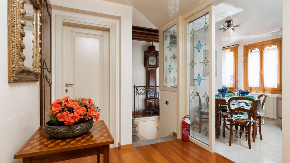 Ingresso interno di un’abitazione con pavimento in legno, tavolo consolle con composizione floreale e specchio dorato, porte bianche e una vetrata decorata che conduce alla zona pranzo. Sullo sfondo si vede una ringhiera in ferro e un orologio a pendolo, mentre la sala da pranzo è illuminata da grandi finestre con infissi in legno.