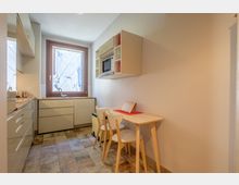 Piccola cucina moderna e luminosa con mobili chiari, lavello sotto una grande finestra e pavimento piastrellato. Sulla parete laterale ci sono un microonde incassato in una mensola e un tavolino in legno con due sedie.