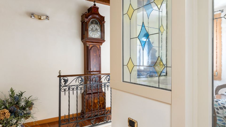 Disimpegno interno con pavimento in legno, ringhiera in ferro battuto e soffitto con travi a vista. Sulla parete è presente un grande orologio a pendolo in legno e una finestra con vetro decorato a rombi colorati.