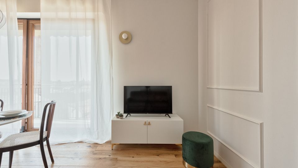 Interno di un soggiorno moderno e luminoso con pavimento in legno, pareti bianche con boiserie e una grande portafinestra con tende leggere. Un mobile TV bianco con televisore e piccola pianta è affiancato da un pouf verde, con tavolo da pranzo e sedie visibili sul lato sinistro.