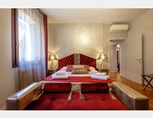 Camera da letto moderna con grande letto matrimoniale al centro, testiera imbottita rossa capitonné e comodini con lampade su entrambi i lati. A sinistra c’è una finestra con tende leggere, mentre a destra si vedono pavimento in legno, porta aperta su un corridoio e un climatizzatore a parete.