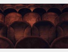 File di poltrone imbottite in velluto marrone, viste in primo piano, disposte in righe parallele come in una sala teatro o cinema. L’illuminazione è soffusa e mette in evidenza le forme arrotondate degli schienali.