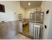 Bagno interno con pareti bianche e rivestimento in piastrelle effetto marmo, pavimento a quadri beige e sanitari sospesi sul fondo. L’area doccia è separata da pannelli decorativi in legno bianco con riquadri a vetro e dettagli dorati, con radiatore bianco in primo piano.