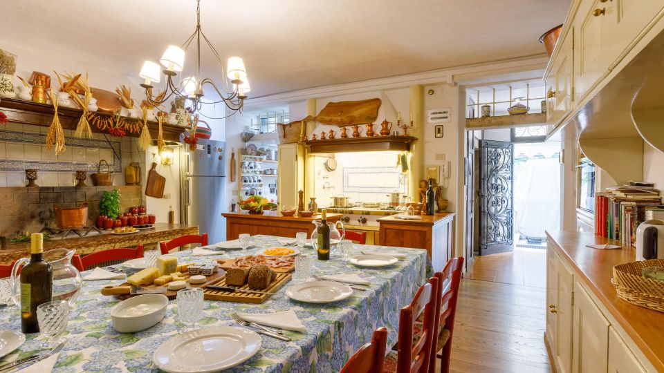 Cucina rustica con grande tavolo da pranzo apparecchiato e sedie rosse, pensili bianchi e piano cottura in muratura con piastrelle decorative. Lampadario a più luci, mensole con utensili e decorazioni in rame, e porta con inferriata che conduce a un piccolo spazio esterno.