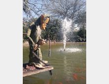 Laghetto in un giardino con una fontana a getto al centro e pesci rossi visibili nell’acqua. In primo piano, su una pedana di legno, c’è una grande statua decorativa di un folletto con cappello e corda, con alberi e siepi sullo sfondo.