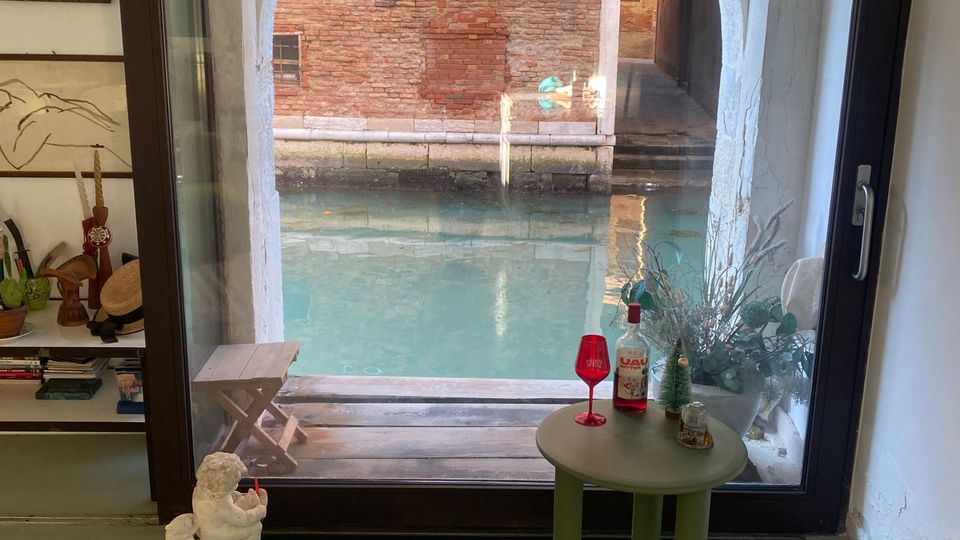 Interno di una stanza con una grande finestra ad arco che si affaccia su un canale veneziano, con un edificio in mattoni visibile all'esterno. La stanza presenta un tavolino verde con oggetti decorativi, un pavimento con un pannello in vetro trasparente e una mensola laterale contenente libri e ornamenti.