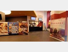 Interno di un cinema con biglietteria “CASSA” sullo sfondo, sportelli con vetri e schermi sopra, pareti in bianco e rosso e pavimento in piastrelle scure. In primo piano sono esposti grandi poster promozionali di film su supporti autoportanti.