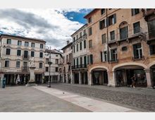 Piazza lastricata in un centro storico, circondata da palazzi di più piani con facciate intonacate, finestre con scuri e balconi in ferro battuto, con portici ad archi al piano terra. Al centro si vedono un monumento su basamento e un lampione decorativo, con cielo parzialmente nuvoloso sullo sfondo.