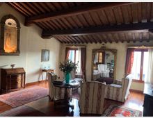 Soggiorno in stile classico con soffitto a travi in legno a vista, pavimento in parquet e tappeti persiani. Al centro ci sono poltrone a righe attorno a un tavolino, una grande specchiera dorata, finestre con tende e mobili antichi lungo le pareti.