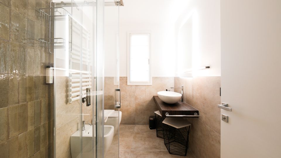 Bagno moderno e luminoso con pareti piastrellate beige e box doccia in vetro a sinistra, sanitari lungo la parete e un lavabo a ciotola su mobile in legno scuro. In fondo c’è una finestra e sul lato destro uno specchio con illuminazione.