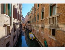Stretto canale urbano tra alti edifici in mattoni e intonaco con finestre ad arco e persiane verdi, affacciati direttamente sull’acqua. Nel canale si vede una piccola barca gialla ormeggiata e, in fondo, un ponticello che attraversa il corso d’acqua.