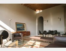 Ampio soggiorno con soffitto alto a travi in legno, pareti chiare e grande arco che conduce alla zona pranzo. Arredi moderni con divani e chaise longue in pelle scura, tavolo con sedie nere, tavolino in vetro e credenza in legno con quadro appoggiato.