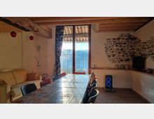 Interno di una sala da pranzo rustica con soffitto in travi di legno, lungo tavolo con sedie e un divano laterale. Una grande porta finestra conduce a un balcone con vista sulle montagne, mentre una parete in pietra a vista e un mobile con TV completano l’ambiente.