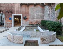 Cortile interno con muri in mattoni e intonaco a vista, un ingresso ad arco e una porta rettangolare che conduce a una galleria. Al centro si trovano strutture in pietra bianca simili a vasche o fontane allineate, circondate da pavimentazione in cemento e siepi verdi lungo il lato destro.