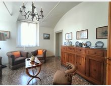 Salotto in stile classico con soffitto a volta e lampadario in ferro battuto, pavimento in graniglia e finestra con tenda chiara. Due poltrone e un tavolino al centro affiancano una lunga credenza in legno con piatti decorativi e quadri appesi alla parete.