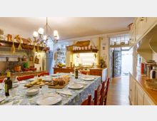 Cucina rustica con grande tavolo da pranzo apparecchiato e sedie rosse, pensili bianchi e piano cottura in muratura con piastrelle decorative. Lampadario a più luci, mensole con utensili e decorazioni in rame, e porta con inferriata che conduce a un piccolo spazio esterno.