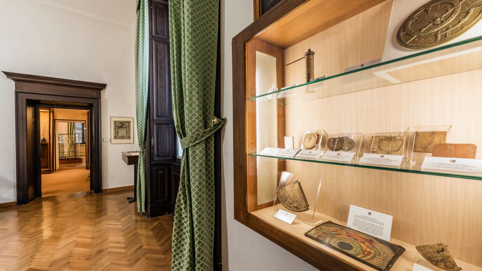Interno di museo con pavimento in parquet a spina di pesce, alte porte in legno scuro e tenda verde accanto a una finestra. A destra, una vetrina a parete con ripiani in vetro espone strumenti e manufatti antichi con cartellini descrittivi.