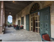 Loggiato di una villa storica con pavimento in pietra e soffitto a travi, pareti affrescate e grandi portoni in legno verde con vetrate. Lungo il corridoio sono disposte poltrone in vimini con cuscini rossi e lampade a sospensione, con arcata aperta verso il paesaggio esterno.