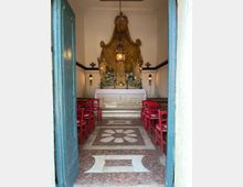 Interno di una piccola cappella con altare centrale decorato da statue e fiori, illuminato da una lampada sospesa e applique laterali. Il pavimento in marmo presenta un motivo geometrico con rosetta centrale e ai lati ci sono file di sedie rosse, visto dalla soglia di una porta azzurra.