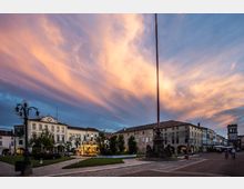 Piazza storica al tramonto con ampi spazi pavimentati e aiuole verdi, circondata da edifici bassi con portici e facciate in stile tradizionale. Al centro si vede un alto pennone su basamento monumentale e, sullo sfondo, una torre con orologio; sono presenti lampioni decorativi e una piccola giostra illuminata.