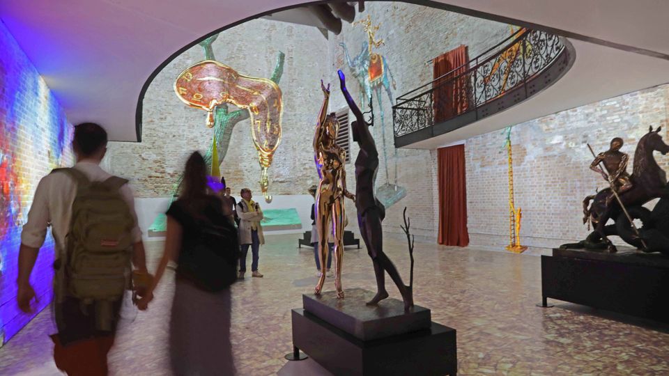 Interno di una galleria d’arte con pareti in mattoni bianchi e soffitto ad arco, con balconata superiore in ferro battuto. Nella sala sono esposte sculture su piedistalli e installazioni sospese, tra cui un grande orologio molle, con tende rosse e pavimento in pietra.