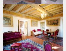 Ampio salone in stile storico con soffitto a travi decorate e lampadario, pareti con grandi affreschi incorniciati e pavimento in cotto con tappeti orientali. Arredi classici in legno con divani e sedie imbottite rosse, tavolo rotondo da pranzo e una porta che conduce a un corridoio adiacente.