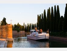 Canale navigabile con una barca bianca in transito tra alti cipressi e argini in pietra e mattoni. A sinistra si vede un muro in pietra con una piccola croce sulla sommità, illuminato dalla luce calda del tramonto.