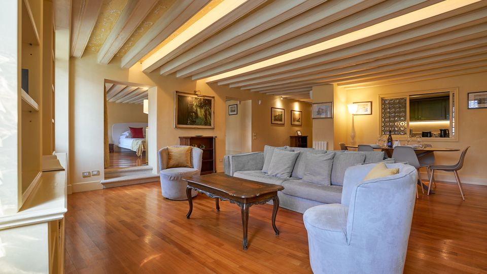 Ampio soggiorno open space con pavimento in legno e soffitto con travi a vista verniciate di bianco, arredato con divano, poltrone e tavolino. Sul fondo si vedono la zona pranzo con cucina a vista e, a sinistra, un’apertura che conduce a una camera da letto rialzata di pochi gradini.