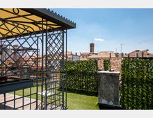 Terrazza panoramica su un tetto con prato sintetico, pergolato in metallo con copertura e siepe verde lungo il parapetto. Sullo sfondo si vedono i tetti in tegole del centro storico e una torre in mattoni sotto un cielo sereno.