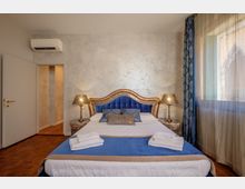 Camera da letto in stile moderno con grande letto matrimoniale al centro, testiera imbottita blu con cornice dorata e due comodini con lampade. Pareti chiare, pavimento in parquet, condizionatore sopra la porta e ampia finestra laterale con tende leggere e drappo blu.