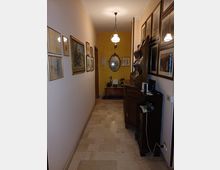 Corridoio interno di una casa con pavimento in pietra chiara, pareti decorate da numerosi quadri incorniciati e lampadario a sospensione. Sul lato destro ci sono mobili in legno con oggetti e una specchiera ovale sulla parete di fondo.