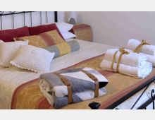 Camera da letto con grande letto matrimoniale in struttura di metallo, copriletto decorato e molti cuscini. Sul letto sono disposte coperte piegate e asciugamani arrotolati legati con nastri, con comodino e lampada sullo sfondo.