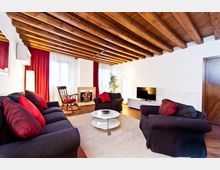 Soggiorno moderno con soffitto a travi di legno a vista, pareti bianche e pavimento in legno. Divani e poltrone scure con cuscini rossi circondano un tavolino rotondo su tappeto chiaro, con TV su mobile basso e grandi finestre con tende rosse e bianche.