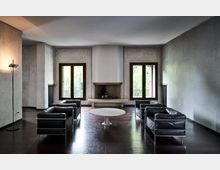 Soggiorno moderno e minimalista con pavimento in legno scuro, caminetto centrale e due grandi finestre con infissi in legno che affacciano sul verde. Al centro ci sono un tavolino rotondo bianco e divani in pelle nera disposti su due lati, con pareti chiare e radiatori ai lati delle finestre.