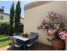 Piccolo giardino con tavolo da esterno e sedie in metallo su un patio, accanto a un muro color crema con bordo merlato. Sono presenti cipressi, prato verde e un grande vaso con arbusto fiorito rosa.