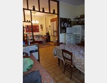 Interno di un appartamento con cucina-soggiorno open space, pavimento in piastrelle e tavolo da pranzo con tovaglia floreale accanto a un frigorifero aperto e mensole a parete. Un separé a scaffali divide la zona cucina dalla zona salotto, dove si vedono divano, quadri, lampadario e una porta-finestra.