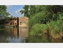 Edifici rurali in muratura, simili a un vecchio mulino, affacciati su un fiume o canale con acqua calma che riflette le facciate. Sulla riva opposta si vedono canneti e vegetazione fitta con alberi alti che incorniciano il corso d’acqua.