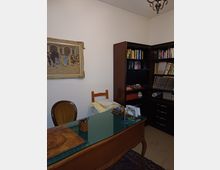 Studio domestico con scrivania in legno e piano in vetro, sedia e blocco appunti, davanti a librerie scure ad angolo piene di libri e raccoglitori. Pareti bianche con quadro incorniciato e lampadario a soffitto, pavimento chiaro con tappeto.