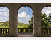 Loggia con tre grandi archi e balaustra in pietra, con pareti affrescate a motivi vegetali. Attraverso le arcate si vede un paesaggio collinare con campi verdi, alberi e cielo parzialmente nuvoloso.