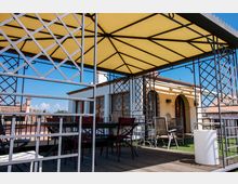 Terrazza panoramica con gazebo in metallo e copertura gialla, pavimento in legno e arredi da esterno come tavoli e sedie. Sullo sfondo si vede una facciata chiara con archi e tetti in tegole sotto un cielo sereno.