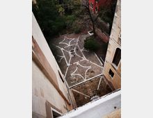 Veduta dall’alto di un cortile lastricato in pietra con un motivo geometrico di linee bianche e un piccolo pozzo centrale. Il cortile è circondato da edifici con facciate intonacate e da alberi e arbusti lungo i lati.
