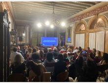 Sala conferenze in un palazzo storico con soffitto a travi dipinte, lampadario centrale e pareti decorate con boiserie e archi con tende bianche. In fondo alla stanza è visibile un grande schermo per presentazioni affiancato da un leggio, con file di sedie rivolte verso lo schermo.