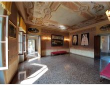 Interno di una sala storica con pavimento in terrazzo e soffitto decorato con stucchi e affreschi, illuminata da finestre e lampade a parete. Le pareti rivestite con motivi damascati espongono quadri e abiti in cornici, con panche imbottite lungo i lati e un corridoio visibile attraverso una porta aperta.