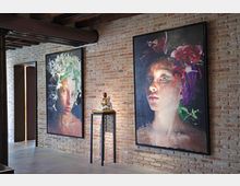 Interno in stile industriale con parete in mattoni a vista, soffitto con travi e condotte, e pavimento in cemento. Due grandi quadri incorniciati appesi alla parete e una piccola scultura su un tavolino alto; a sinistra si vede un corridoio con illuminazione verticale.